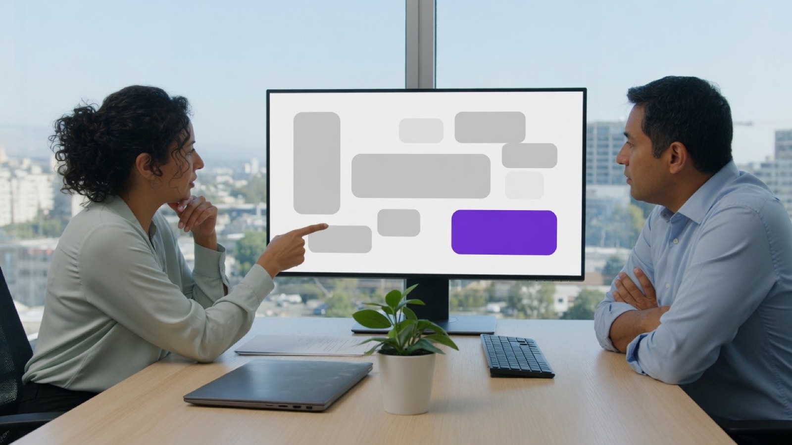 Two people reviewing a layout of gray blocks and a violet button on a monitor.